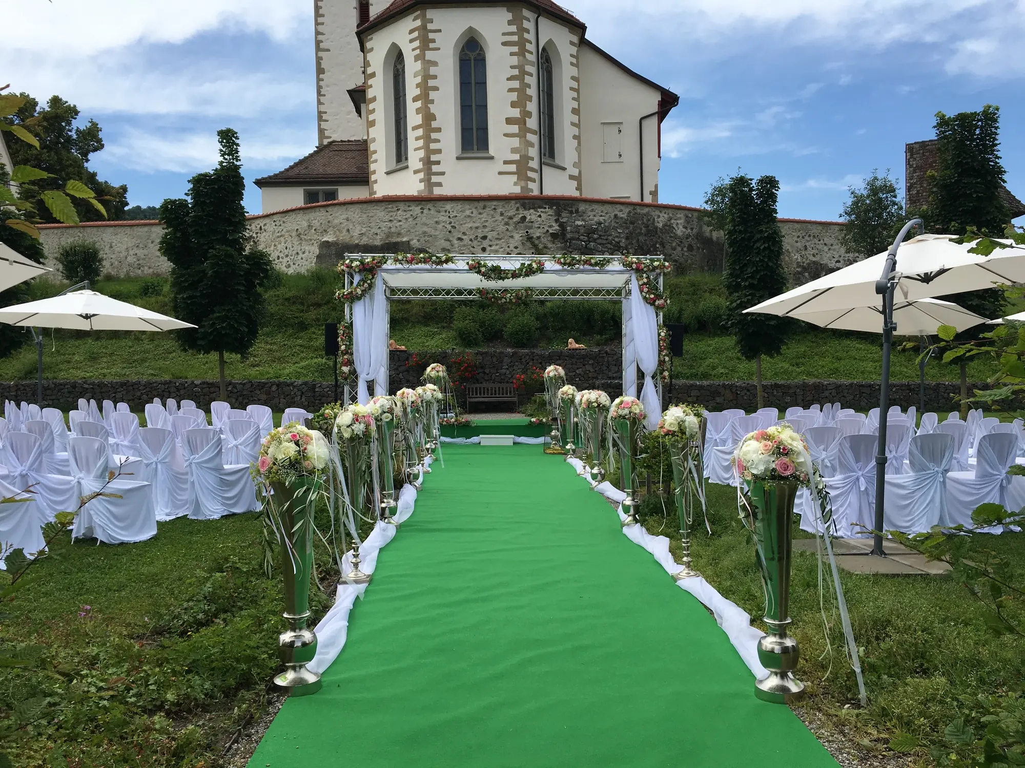 Freie Trauung im Garten mit grünem Teppich, Blumenschmuck, Traubogen und Kirche im Hintergrund